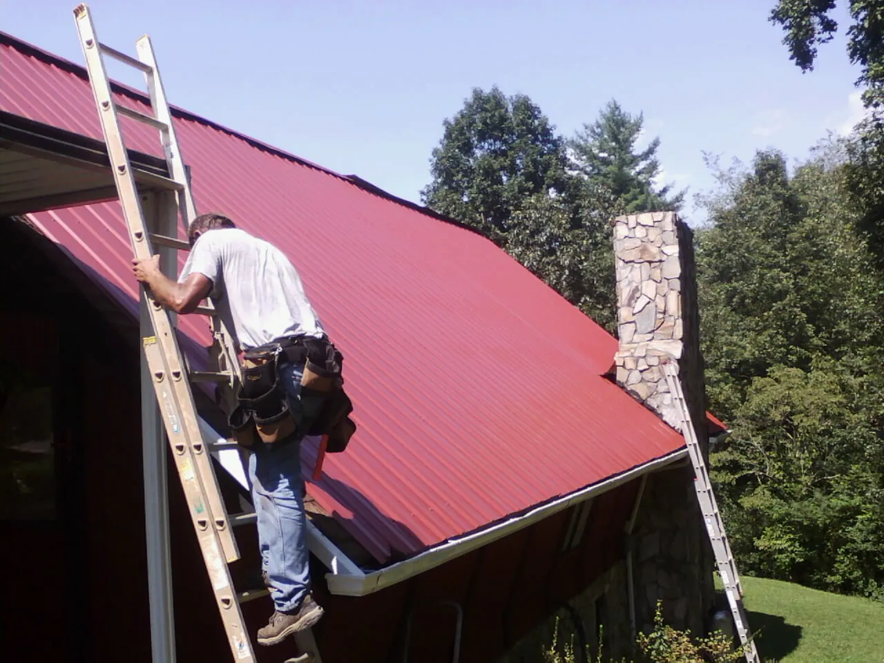 Roofer on a ladder beside a red metal roof and stone chimney — Soffit & Fascia Repair by Roofing Brothers in Vandiver